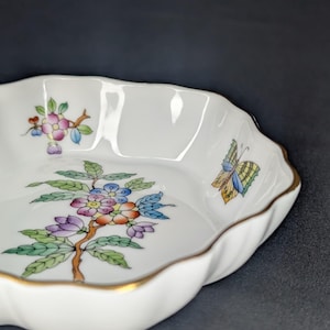 May include: White porcelain dish with a scalloped edge and gold trim. The dish features a colorful floral and butterfly design. The flowers are pink, purple, and blue, with green leaves. A decorative piece.