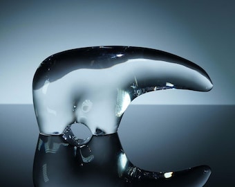 Op de afbeelding: Een heldere glazen sculptuur met een gebogen, snavelachtige vorm. Het object is zeer reflecterend, met een glad oppervlak en een cirkelvormige opening. De sculptuur staat op een reflecterend oppervlak, waardoor een spiegeleffect ontstaat. De achtergrond is een verloop van blauw en grijs.