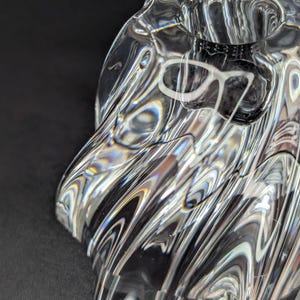 Pode incluir: Uma tigela decorativa de vidro transparente com um design ondulado e texturizado. A tigela tem uma abertura circular e é feita de vidro transparente, refletindo a luz e criando um jogo de cores. O design da tigela lembra água corrente.
