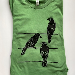Op de afbeelding: Een opgevouwen groen t-shirt met een zwarte afbeelding van drie kraaien op horizontale lijnen. De kraaien hebben gedetailleerde zwarte markeringen. Het shirt is gemaakt van een zacht materiaal.
