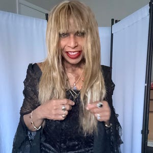May include: A black lace long-sleeve top with a textured pattern, worn with layered silver-tone necklaces featuring blue and red pendants, a silver-tone bracelet, and two silver-tone rings with dark stones on the fingers of each hand.