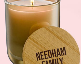 Puede incluir: Una vela encendida en un frasco de vidrio ámbar translúcido con una tapa redonda de bambú. La tapa presenta el texto marrón oscuro NEEDHAM FAMILY impreso en una fuente sans-serif. La cera de la vela es de un color tostado claro y cálido.