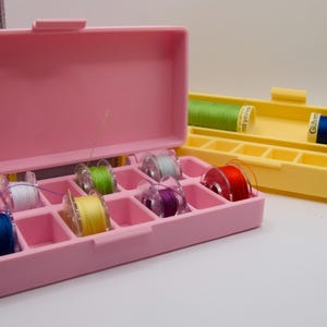 Puede incluir: Cajas de almacenamiento de hilo de plástico rosa y amarillo. La caja rosa está abierta, revelando carretes de hilo colorido. La caja amarilla está abierta, mostrando dos carretes de hilo más grandes. Suministros de costura y artesanía.