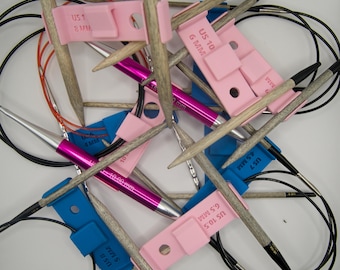 May include: A collection of knitting needles and accessories. The image shows wooden needles, pink and blue needle size markers, and black cables. The markers are labeled with US sizes and mm measurements. The needles are scattered across a white surface.