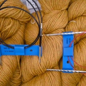 May include: Close-up of knitting needles with blue connectors and black and red cables. The blue connectors have the text "USA 4 3.5 MM" and "US 6 4MM". The needles are set against a background of golden-brown yarn.