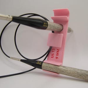 以下が含まれることがあります： ピンク色の針ホルダーが付いた編み針のクローズアップ。針は木製で、黒い先端と黒いケーブルが付いています。ピンク色のホルダーには「US 13 9 MM」と書かれています。
