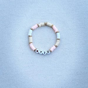 May include: A beaded bracelet with a mix of pink, white, and tan disc-shaped beads. The bracelet includes gold-colored spacers and white letter beads spelling "Y2D" with heart accents. The bracelet is on a light blue textured surface.