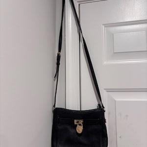 May include: Black leather crossbody bag with a long adjustable strap. The bag features a gold-tone lock closure and hardware. The bag is hanging on a white door.