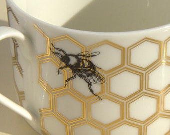 Może przedstawiać: Biały ceramiczny kubek do herbaty z wzorem w kształcie plastra miodu w kolorze złotym i czarnym rysunkiem pszczoły.
