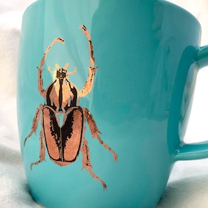 Könnte beinhalten: Eine türkisfarbene Keramiktasse mit einem goldenen Käferdesign. Der Käfer hat einen braunen Körper mit schwarzen Markierungen.