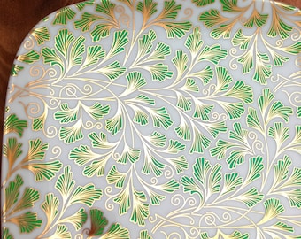 May include: A white ceramic plate with a green and gold floral pattern. The pattern features stylised leaves and swirls.