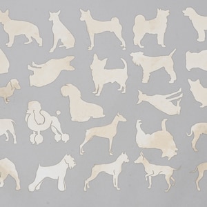 Peut inclure: Une collection de 20 silhouettes de races de chiens différentes découpées dans du bois. Les silhouettes sont peintes dans une couleur beige clair et sont disposées de manière aléatoire sur un fond gris.