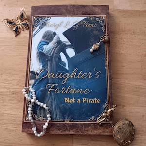 May include: A book titled "Daughter's Fortune: Not a Pirate" with a cover illustration of a person at a ship's wheel. A gold butterfly brooch, a pearl necklace, and a gold pendant with a decorative design are placed around the book.