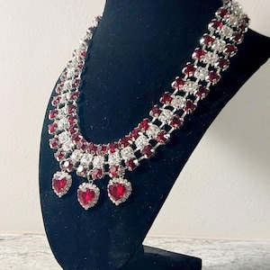 May include: A sparkling necklace with alternating rows of red and clear crystals. Three heart-shaped pendants with red crystals dangle from the necklace. The jewelry is displayed on a black mannequin.