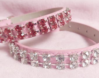 Puede incluir: Dos collares rosas para mascotas con filas de brillantes pedrería. Un collar presenta pedrería roja, mientras que el otro tiene pedrería transparente. Los collares están hechos de un material rosa claro y están diseñados para mascotas pequeñas.