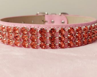 Puede incluir: Collar rosa para mascotas con tres filas de brillantes pedrería rojo-anaranjada. El collar tiene una hebilla plateada y está sobre un fondo rosa suave. Un accesorio elegante para una mascota.