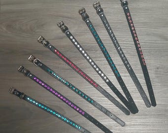 May include: A collection of black pet collars with silver buckles and decorative rhinestones in various colors, including purple, teal, white, and red. The collars are arranged diagonally on a wooden surface.