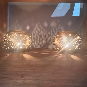 May include: Two silver candle holders with a geometric pattern, illuminated by candles, casting shadows on a wooden surface.