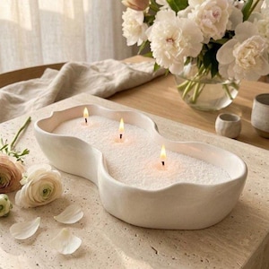 Puede incluir: Un portavelas blanco en forma de onda con tres mechas encendidas. El portavelas está lleno de cera blanca y se encuentra sobre una superficie clara. Pétalos de flores blancas y un jarrón con flores blancas en el fondo.