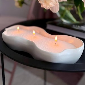 Op de afbeelding: Een witte, golvende kaarsenhouder met drie brandende lonten. De houder is gevuld met witte was en staat op een zwarte tafel. De kaars is een decoratief woonaccessoire.