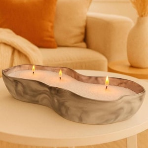May include: A large, grey, three-wick candle in a wavy, oblong container. The candle is lit, with three flames visible. The container has a marbled appearance and sits on a white table. The background includes a sofa and decorative items.
