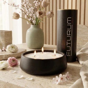 May include: A black, low-profile, round candle bowl with three lit wicks and white wax, accompanied by a tall black cylindrical container labeled FUTURUM in white vertical text, set near a light green vase with light pink flowers.