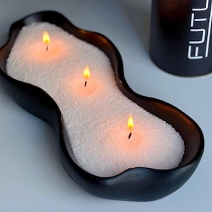 Pode incluir: Um castiçal preto com bordas onduladas e três velas acesas. As velas estão em grânulos brancos e as chamas são amarelo brilhante. Um objeto cilíndrico preto com texto branco está no fundo.