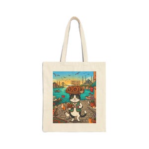 May include: A beige canvas tote bag featuring a colorful illustration of a black and white cat wearing a green vest, balancing a tray of simit bread on its head, with a background of a coastal city, boats, and the text Simitçi Kedi Günlük ve Taze.