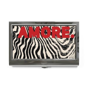 May include: A rectangular metal business card holder with a polished silver finish. The front panel features a cream background with a black wavy zebra-like pattern and the word AMORE. printed in bold red capital letters across the center.