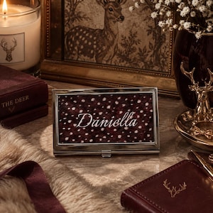 May include: A rectangular silver-toned metal business card holder featuring a dark brown and white spotted faux fur inlay on the front cover. The name Daniella is engraved in a white cursive script across the center of the spotted pattern.