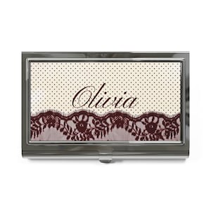May include: A rectangular metal business card case featuring a cream-coloured background with small black polka dots. The lower portion displays a dark brown lace trim pattern. The name Olivia is printed in a dark brown cursive script in the centre.