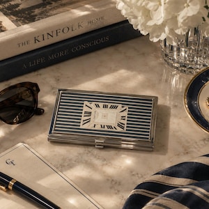 May include: A rectangular metal business card case featuring a navy blue and white striped pattern on the exterior. The center of the case displays a white rectangular inlay with black Roman numerals and the Cartier brand name printed in black.
