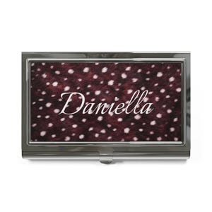 May include: A rectangular metal business card case with a polished silver finish. The center panel features a dark maroon background with a white spotted pattern and the name Daniella printed in a white cursive script font across the middle.