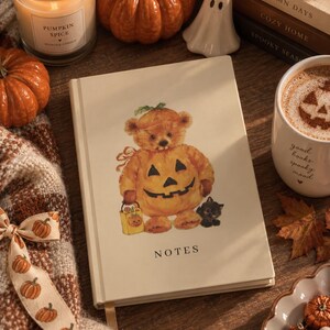 Puede incluir: Un cuaderno de tapa dura de color crema con la palabra NOTES impresa en negro en la parte inferior. La portada presenta una ilustración central de un oso de peluche marrón con un disfraz de calabaza, sosteniendo una pequeña bolsa amarilla y un gato negro.