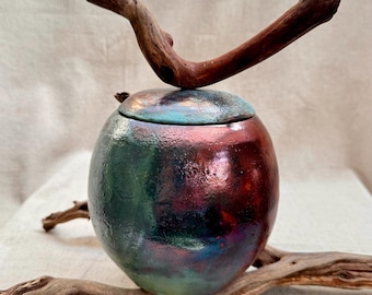 Op de afbeelding: Een ronde keramische pot met een iriserende groene, blauwe en rode glazuur. De pot heeft een deksel en staat op een stuk drijfhout.