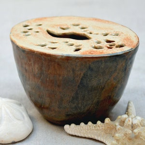 May include: A ceramic planter with a light brown glaze and a cutout design. The planter has a rustic look and is perfect for displaying small plants or flowers.