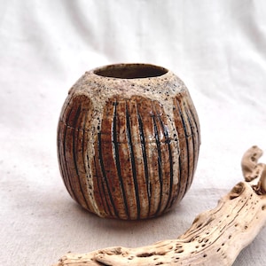 May include: A round ceramic vase with a textured brown and tan surface featuring vertical carved lines. The upper rim has a speckled cream-colored glaze that drips down onto the darker brown body. The vessel has a wide, bulbous shape.