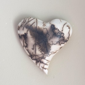 May include: A heart-shaped ceramic piece with a white base and intricate dark brown patterns. The design resembles a map or abstract artwork, with a central dark area and delicate lines.