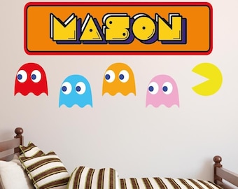 Op de afbeelding: Een muursticker met de naam "MASON" in een retro-videogamestijl, met vier Pac-Man-geesten in rood, blauw, oranje en roze, en een gele Pac-Man.
