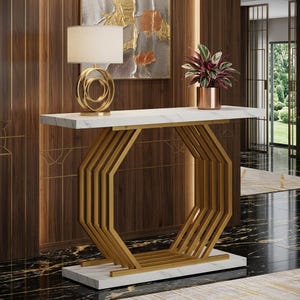 May include: A console table featuring a white marble top and base with a gold-toned metal frame composed of repeating octagonal shapes. A table lamp with a gold circular base and a white drum shade sits on the left, and a potted plant sits on the right.