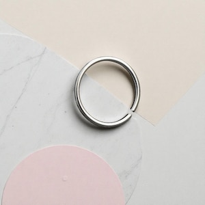 Puede incluir: Un anillo de aro abierto de color plateado. El anillo es circular con un pequeño espacio y se encuentra sobre un fondo de tonos neutros, que incluyen formas geométricas blancas, beige y rosas. El anillo es de diseño simple y minimalista.