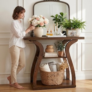 Puede incluir: Una mesa consola de madera oscura con marco curvo, que presenta un estante superior con un jarrón blanco de flores, difusores de varillas, velas y un marco de fotos. El estante inferior contiene libros apilados, una planta y una cesta tejida.