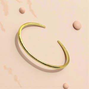 May include: A gold-colored cuff bracelet with a simple, open design. The bracelet has a smooth, polished finish and is set against a light pink background with three small, round, pink objects. The jewelry is a minimalist accessory.