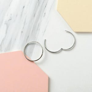 May include: Two silver hoop earrings, one complete and one open, are displayed on a white marble surface with geometric shapes in pink and beige. The earrings have a simple, minimalist design.