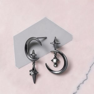 Puede incluir: Un par de pendientes grises oscuros con un diseño de luna creciente y estrella. Cada pendiente tiene forma de media luna con detalles de estrella. Un pendiente tiene una estrella colgante con una gema transparente, y el otro tiene una estrella con una gema transparente y una gema colgante.