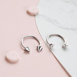 Puede incluir: Dos pendientes plateados en forma de herradura. Un pendiente tiene extremos puntiagudos y el otro tiene extremos claros, redondos y con forma de gema. Los pendientes están sobre un fondo rosa y blanco con dos objetos rosas pequeños y redondos.