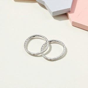 May include: Two silver hoop earrings with a textured, etched design. The earrings are circular and have a shiny, metallic finish. The background is a light, neutral color.