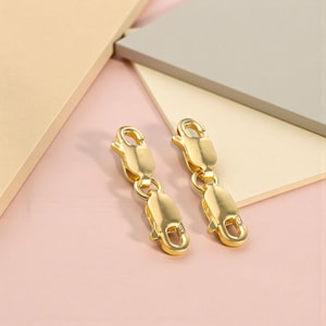 Puede incluir: Dos cierres de joyería dorados con diseño de mosquetón. Cada cierre tiene un pequeño lazo redondo y un cuerpo rectangular. Los cierres están conectados por un pequeño anillo. El fondo es una mezcla de formas geométricas rosas, beis y grises.