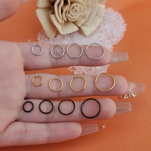 May include: A variety of hoop earrings in different sizes and colors, including silver, gold, and black. The hoops are displayed on a hand, with the smallest hoop at the top and the largest hoop at the bottom.