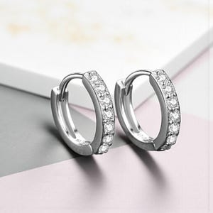 May include: A pair of silver hoop earrings. Each earring is set with small, clear gemstones along the outer curve. The earrings are displayed on a white and pink surface.